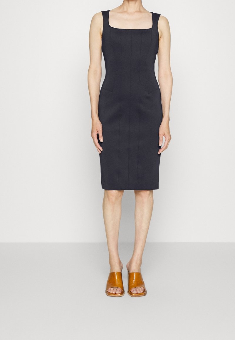 Femme portant une robe noire ajustée sans manches, longueur genou, avec un décolleté carré, et des mules marron à bout ouvert, se tenant devant un fond blanc.