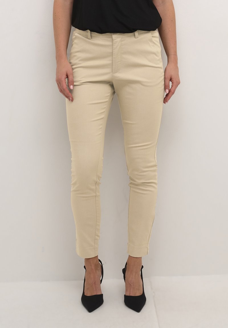 Personne portant un pantalon beige ajusté et des chaussures à talons noires, debout devant un fond clair uni.
