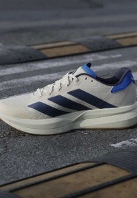 Chaussure de running blanche avec des bandes bleu marine et des accents bleus sur l'asphalte près de dos d'âne jaunes et noirs.