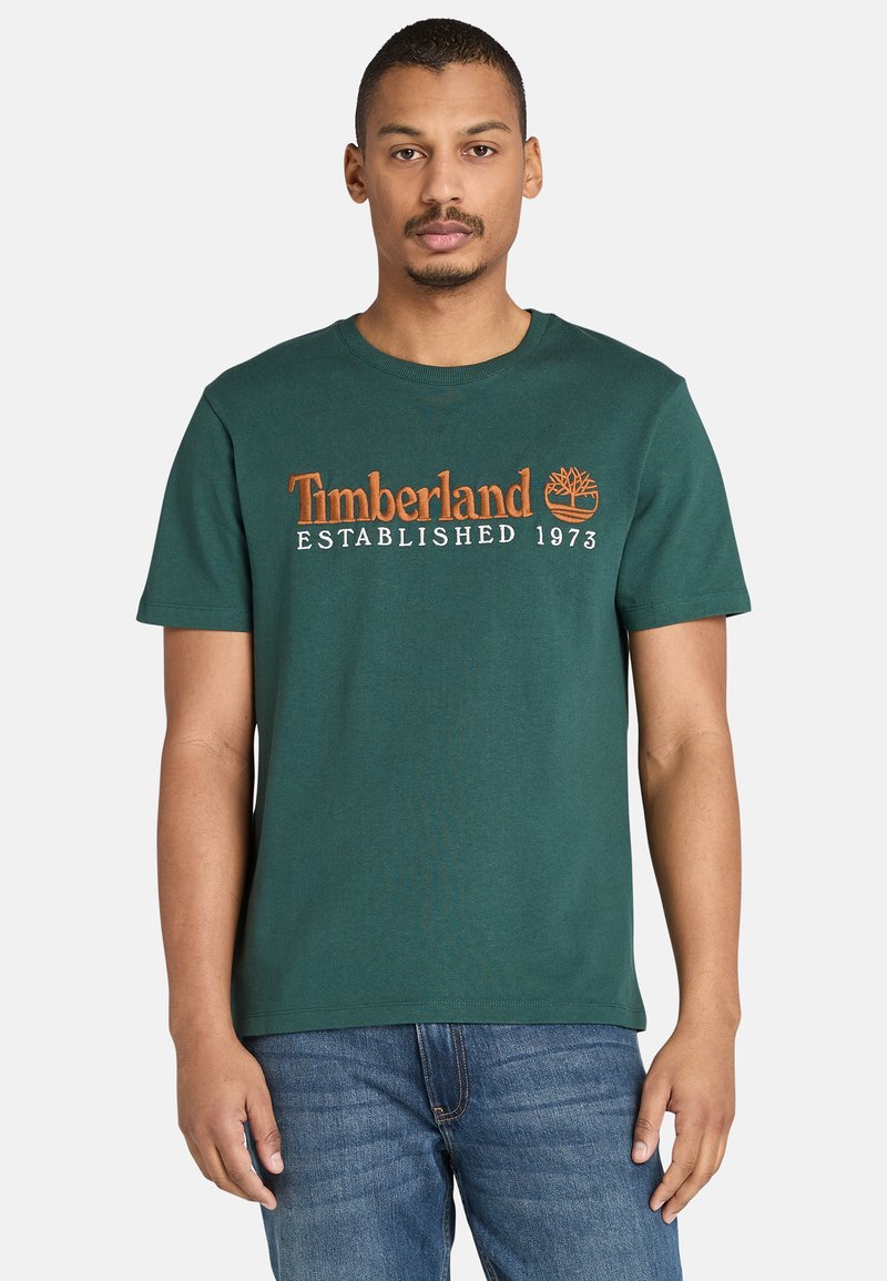 Muž ve zeleném tričku Timberland s oranžovým logem a textem, v kombinaci s modrými džíny, stojící proti jednoduchému světlem pozadí.