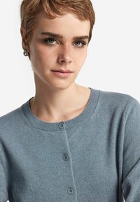 Jasnoniebieski kardigan, klasyczny okrągły dekolt, zapięcie na guziki, ściągacze na rękawach. Miękka tekstura, lekki materiał o delikatnym połysku.