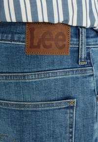Jeans de denim azul com um patch de couro castanho exibindo o nome da marca "Lee", apresentando costuras douradas e bolsos traseiros com costuras detalhadas.