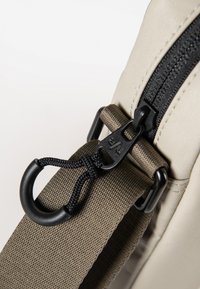 Dettaglio di una borsa beige con una cerniera nera, accessori neri e una tracolla testurizzata grigio scuro con un motivo intrecciato e clip attaccata.