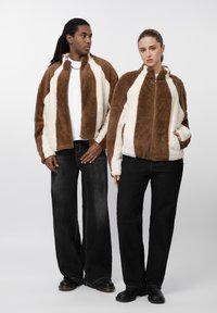 Brune og cremefarvede, fluffy jakker med lynlåslukning foran og stribet design. Begge modeller har sorte jeans og mørke sko på og poserer foran en neutral baggrund.