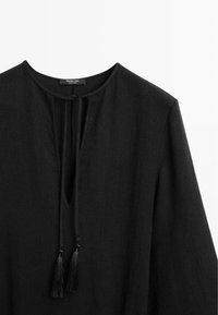 Blouse noire à manches longues avec encolure ronde, fente en V à l'avant avec liens à pompons, et tissu texturé.