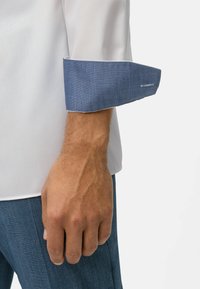 Camicia bianca con un polsino blu texturizzato, con bordi blu navy. Mano visibile con una struttura venosa definita, che indossa pantaloni a motivi blu scuro.