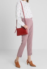 Rød håndtaske med krokodilletekstur og lang rem, båret med en hvid bluse med farverige prikker og lyserøde, smalle bukser.