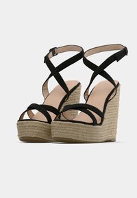 Black strappy wedge sandals with an jute rope platform. The straps are made of soft material, featuring a crossed design over the foot.
