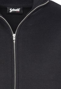 Sort zip-up jakke lavet af tekstureret stof, med høj krave og sølv lynlås. Logoet er synligt ved halsudskæringen.