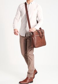 Bolso mensajero de cuero marrón con textura suave, doble asa superior y correa larga, con dos bolsillos delanteros y detalles metálicos mínimos.