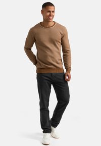 Brązowy sweter z prążkowanymi mankietami i dołem, okrągłym dekoltem, w połączeniu z ciemnymi dżinsami i białymi sneakersami. Modelka stoi na neutralnym tle.