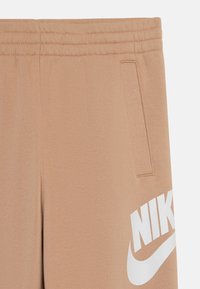 Beige krāsas Nike sporta bikses ar gludu tekstūru, elastīgu jostasvietu, sānu kabatu un baltu logotipa apdruku uz apakšējā kājas daļas.