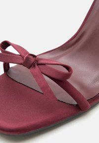 Gros plan sur une sandale bordeaux à bout carré avec un nœud décoratif en fines lanières de tissu sur le devant.