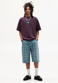 Lilla oversized t-shirt med grafisk detalje, lyseblå denim shorts med gentaget logomønster, hvide crew socks og sorte sko.