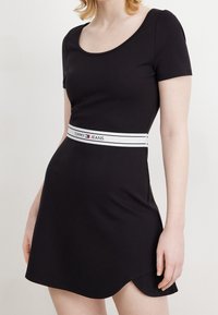 Nainen, joka pukeutunut mustaan lyhythihaiseen mekkoon, jossa on valkoinen vyötärönauha, johon on merkitty "Tommy Jeans", yksinkertaista valkoista taustaa vasten.