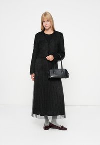 Cardigan noir en peluche, robe longue noire texturée avec une couche transparente, chaussettes grises, chaussures bordeaux brillantes et un petit sac à main noir.