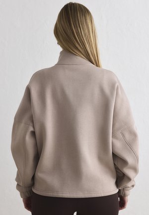 Vrouw met lang blond haar, een loszittende beige sweater dragend, staand met haar rug naar de camera tegen een effen lichte achtergrond.