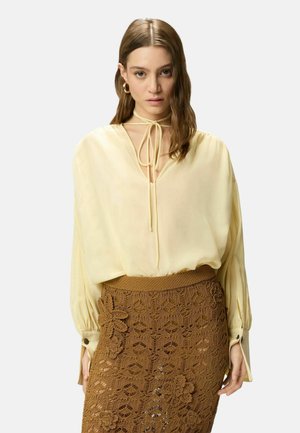 Giovane donna che indossa una camicetta crema larga con scollo a laccio e una gonna marrone floreale all'uncinetto, in piedi davanti a uno sfondo semplice.