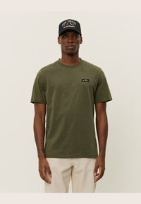 Olive green cotton t-shirt with short sleeves and a crew neckline, featuring a black rectangular logo patch on the left chest.