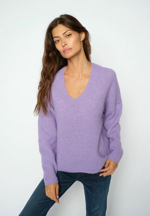 Frau mit langen braunen Haaren, die einen lavendelfarbenen V-Ausschnitt-Pullover und dunkelblaue Jeans trägt, steht vor einem schlichten weißen Hintergrund.