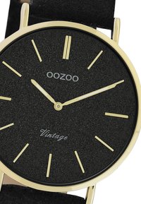 Cadran de montre noir texturé avec des index et aiguilles dorés. Boîtier doré, bracelet noir. Le texte indique "OOZOO" et "Vintage" en argent.
