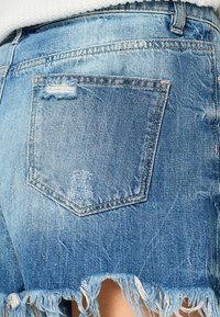 Niebieskie szorty dżinsowe z postrzępionymi brzegami, z tylną kieszenią o postarzanym wykończeniu i jasnym odcieniu, podkreślające elementy designu codziennego stylu.