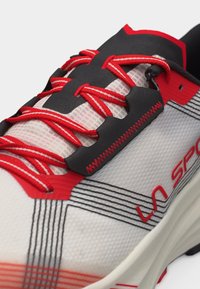 Κοντινό πλάνο λευκού αθλητικού παπουτσιού με κόκκινες λεπτομέρειες, κόκκινες και μαύρες κορδόνια, μαύρα ράμματα και μερικό κόκκινο λογότυπο «La Sportiva» στην πλευρά.