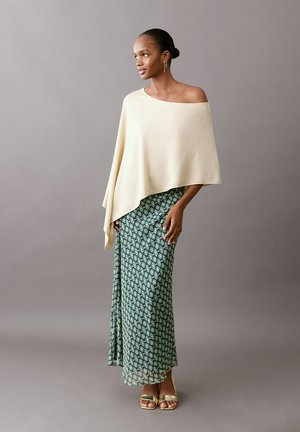 Femme debout portant une jupe verte à motifs, un pull crème asymétrique à épaule dénudée, des sandales dorées et des boucles d'oreilles pendantes dorées sur fond gris.