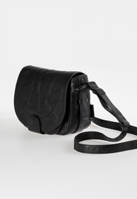 Musta nahkainen crossbody-laukku, jossa on pyöreä läppä, säädettävä olkahihna ja sileä pinta. Varustettu etulähellä ja minimalistisella muotoilulla.