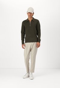 Dark green zippered sweater, long sleeves, with beige pleated trousers and white sneakers. A light-colored cap completes the outfit.