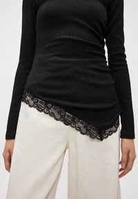 Schwarzes langärmliges Oberteil mit strukturierter Oberfläche und einem zackigen Spitzenabschluss. Kombiniert mit hellen, weit geschnittenen Hosen.