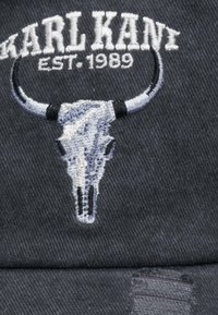 Fekete textília, kiemelkedő szürke hímzett bika koponya mintával, ívelt szarvakkal és a "KARL KANI" felirattal felette, 1989-es alapítással.