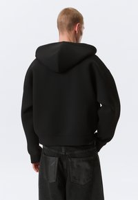 Homme aux cheveux blonds courts portant un sweat à capuche noir ample et un jean sombre avec des poches arrière en cuir, vu de dos sur un fond uni.