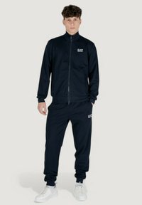 Tuta blu navy realizzata in tessuto liscio, con giacca zip e pantaloni coordinati, entrambi con accenti di logo discreti. Scarpe da ginnastica bianche completano il look.