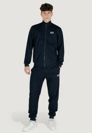 Tuta blu navy realizzata in tessuto liscio, con giacca zip e pantaloni coordinati, entrambi con accenti di logo discreti. Scarpe da ginnastica bianche completano il look.
