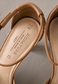 Sandalias de cuero color tan con textura suave, que cuentan con una plantilla acolchada y una correa ajustable. Detalles de talla y marca grabados en la plantilla.