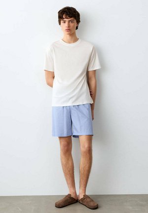 Mladý muž stojaci opretý o bielu stenu, oblečený v bielom tričku, svetlomodrých pruhovaných šortkách a hnedých slip-on topánkach.