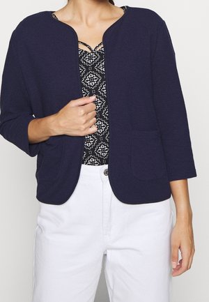 Cardigan texturé bleu marine foncé avec des manches trois-quarts, un col rond et deux poches avant, superposé sur un haut à motifs noir et blanc.