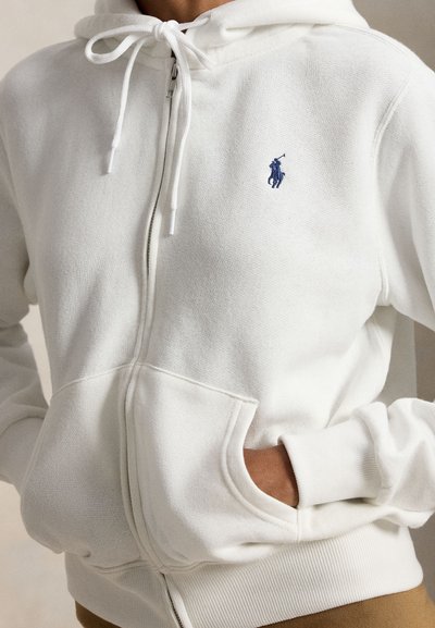Person wearing a white zip-up hoodie with drawstrings and a navy blue embroidered polo player logo, hands in front pockets.