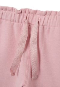 Un pantalon de jogging rose présente une taille élastique avec un cordon de serrage, orné d'accents métalliques. Texture douce et lisse sur tout le tissu.