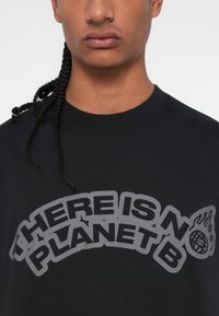 Melna kokvilnas t-kreklu ar lielu pelēku drukātu saukli "NAV CITA PLĀNTA B" ar liesmu un globusa grafiku kā akcentu.