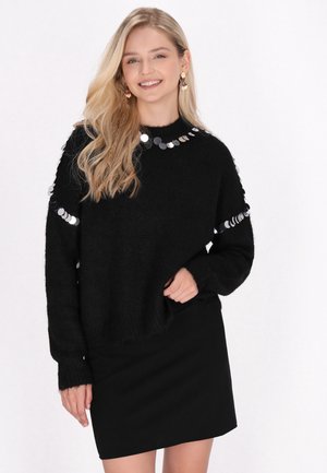 Donna sorridente con lunghi capelli biondi indossa un maglione nero con paillettes argento e una gonna nera su uno sfondo chiaro.
