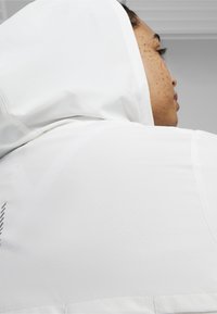 Vit huva jacka med en texturerad yta, med ett triangulärt mönster och en sömdetalj på axeln.