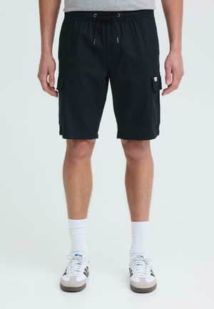 Hombre vistiendo pantalones cortos cargo negros con cinturilla con cordón, calcetines blancos y zapatillas blancas y negras, de pie contra un fondo blanco.