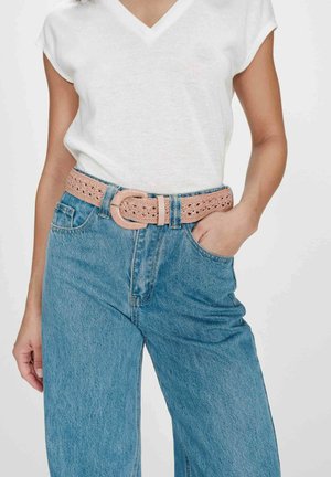 Femme portant un jean bleu clair avec une ceinture beige tissée et un haut blanc à manches courtes avec encolure en V, main dans la poche.