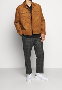 Brun mocka jacka, svart skjorta, grå rutig byxor, vita sneakers med gröna detaljer. Jackan har tryckknappar och bröstfickor.