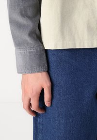 Bluză cu mânecă lungă, cu dungi crem și gri, având manșete contrastante, asortată cu pantaloni din denim albastru. Prim-plan al unei mâini odihnindu-se pe țesătura de denim.
