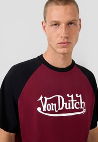 Bordo ir juodas raglan stiliaus marškinėliai, pagaminti iš medvilnės, su ryškiu baltu Von Dutch logotipu priekyje. Minkšta tekstūra, laisvalaikio dizainas.