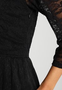 Musta pitsimekko, jossa on läpinäkyvät kuviopaidat ja laskostettu hameosa, esittelee henkilön ylävartalon ja koukistuneen käden.