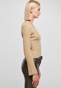 Beige bordázott kötött felső mély V-nyakkal és haranggal végződő hosszú ujjakkal; fényes, sötétbarna, testhezálló nadrággal párosítva. Sima textúra.